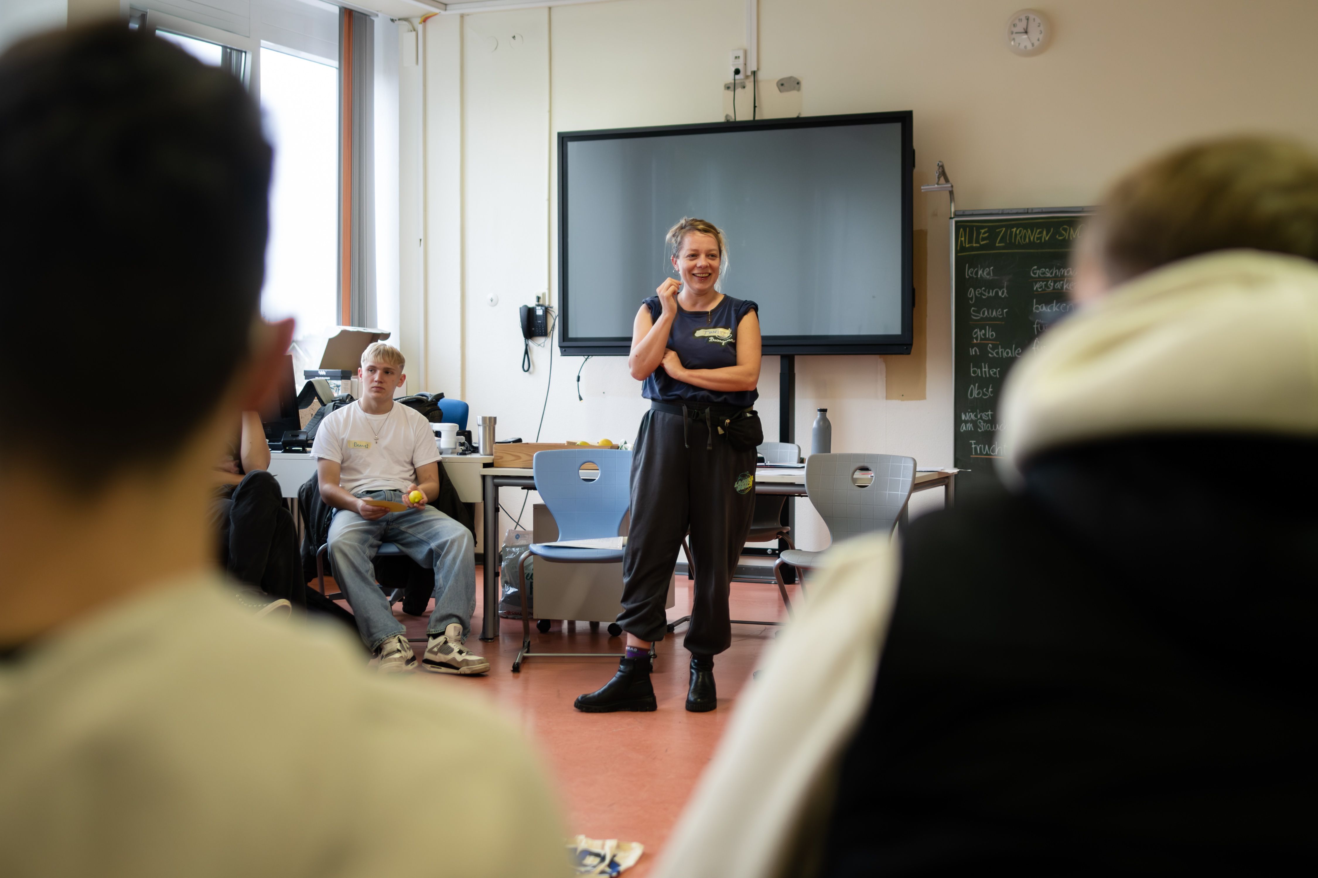 Szene in einer Berufsschulklasse, in der Bildmitte steht eine Bildungsreferentin, die mit den Schüler*innen interagiert. Die Personen im Bild sind weiß gelesen.