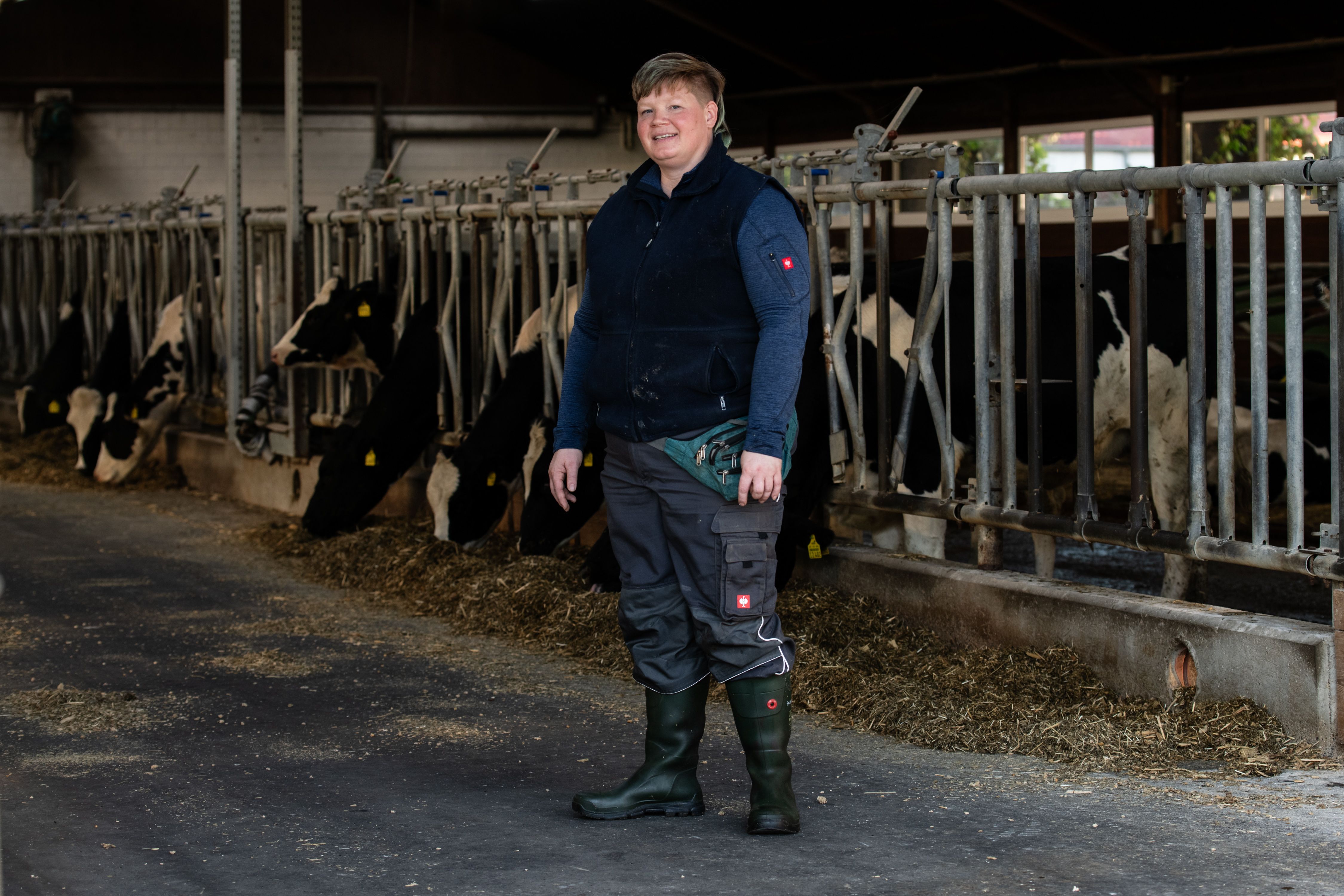 Frau in Gummistiefel steht mit dem Blick in die Kamera. Im Hintergrund ist ein Boxenlaufstall mit Kühen zu sehen. 