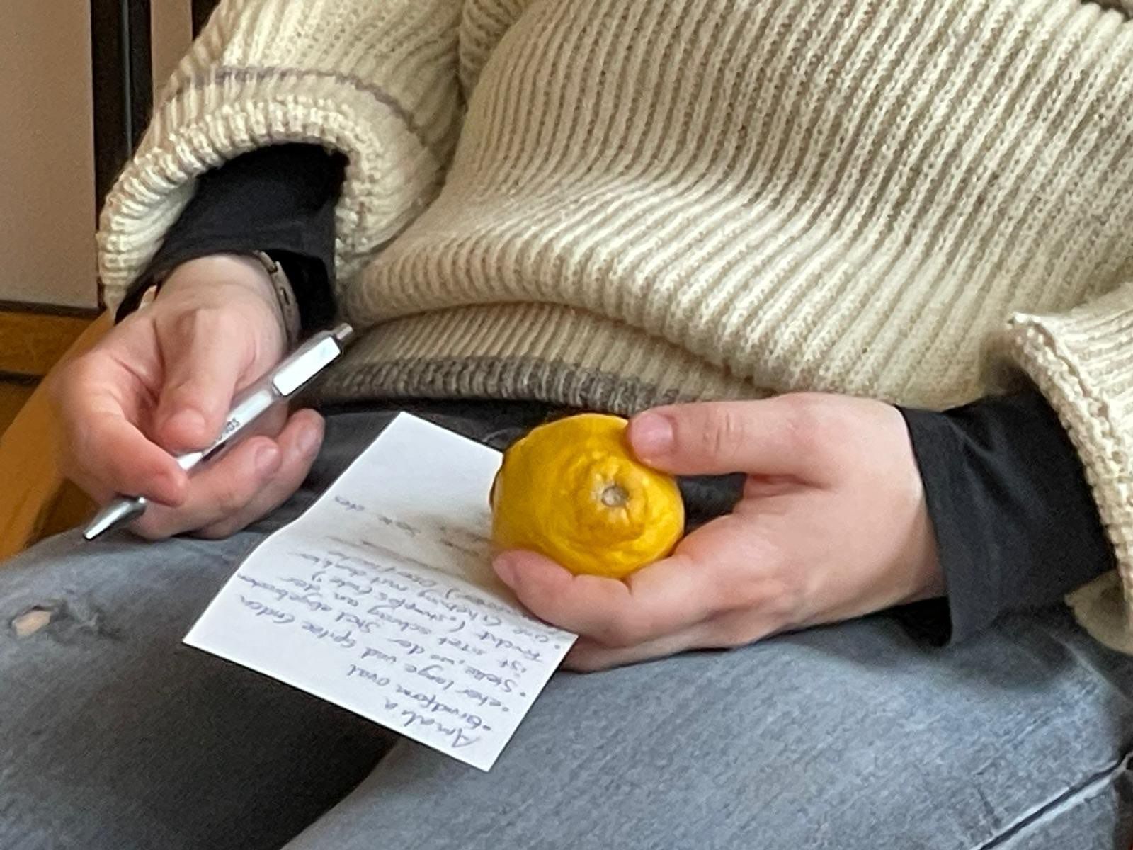 Bildausschnitt einer weißen Person die sitzt und in der linken Hand eine Zitrone hält, in der rechten Hand einen Stift.